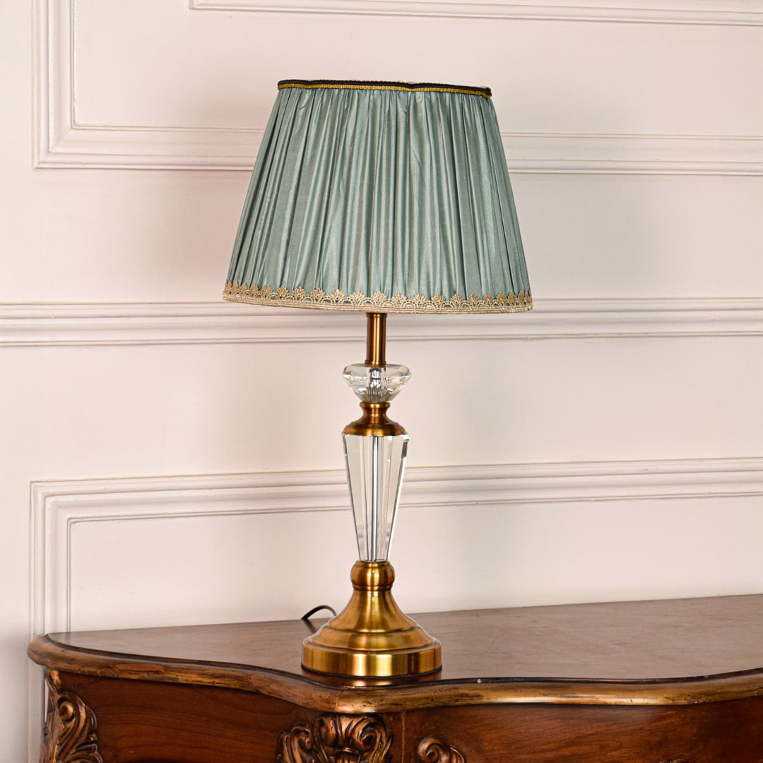 Decorative glass table lamp with gold metal base and pleated teal fabric shade placed on soft off white background.