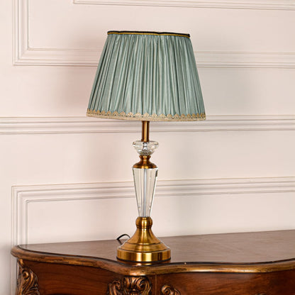 Decorative glass table lamp with gold metal base and pleated teal fabric shade placed on soft off white background.