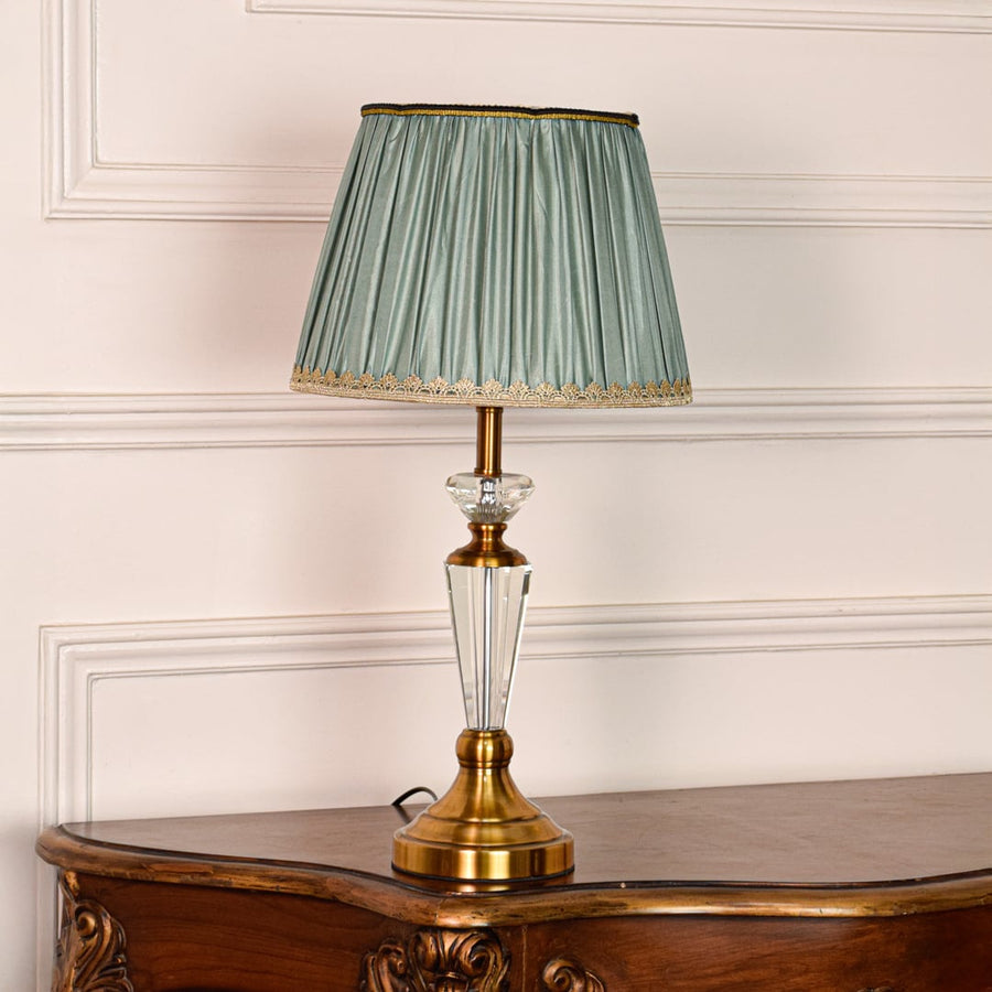 Decorative glass table lamp with gold metal base and pleated teal fabric shade placed on soft off white background.