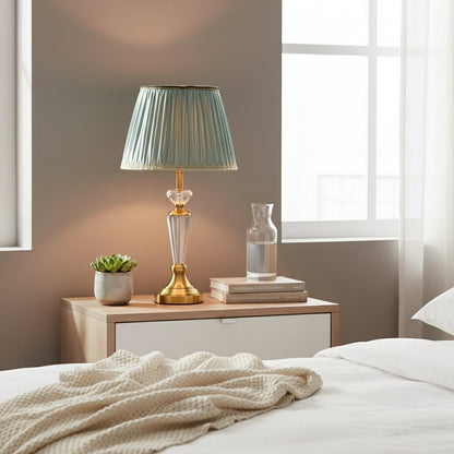 Classic table lamp with clear glass stem gold metal base and pleated teal fabric shade styled on elegant bedside table.