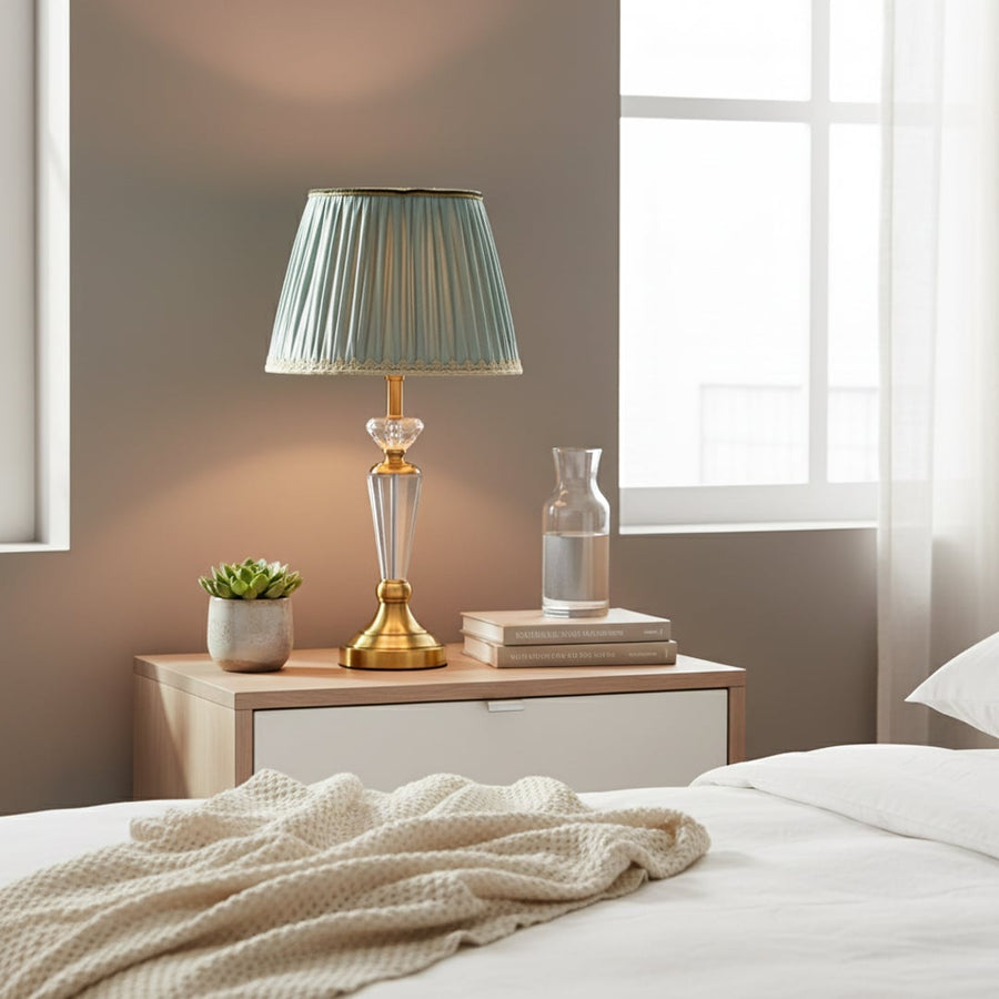 Classic table lamp with clear glass stem gold metal base and pleated teal fabric shade styled on elegant bedside table.