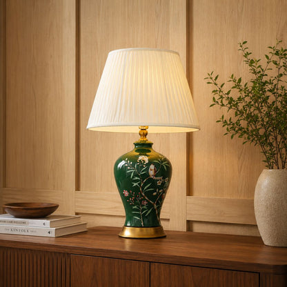 A decorative table lamp with a green ceramic base featuring white floral and bird motifs, a white pleated drum shade, and gold-toned metal accents, shown lit on a wooden console in a warm interior setting.