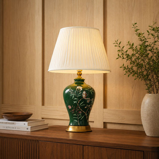 A decorative table lamp with a green ceramic base featuring white floral and bird motifs, a white pleated drum shade, and gold-toned metal accents, shown lit on a wooden console in a warm interior setting.