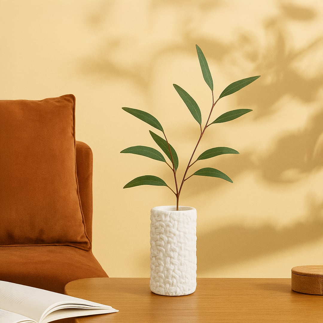 White textured cylindrical ceramic vase with green leaf stems placed on a wooden table beside a rust-colored cushion.