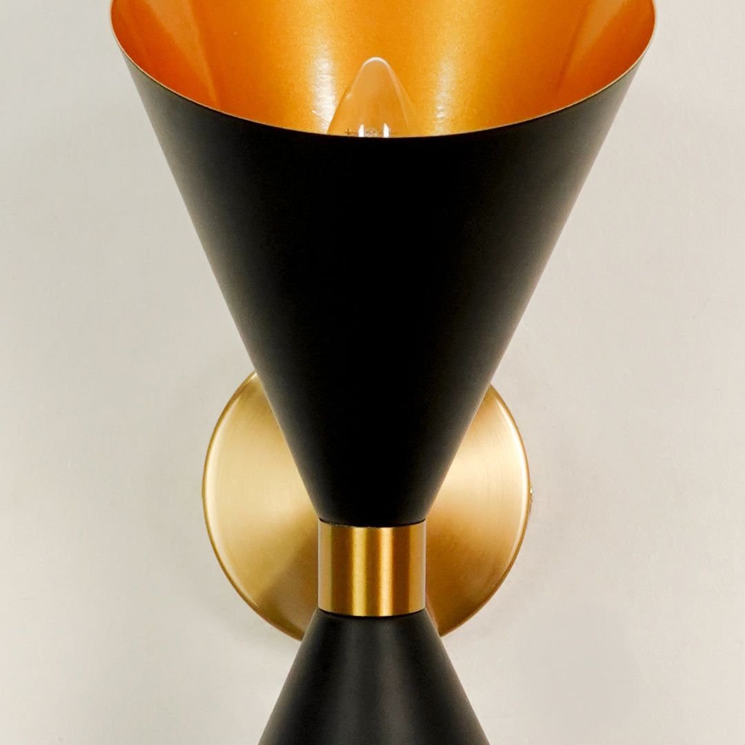 Close up details of an illuminated black and gold brass hourglass wall sconce showcasing the internal golden cone lining.