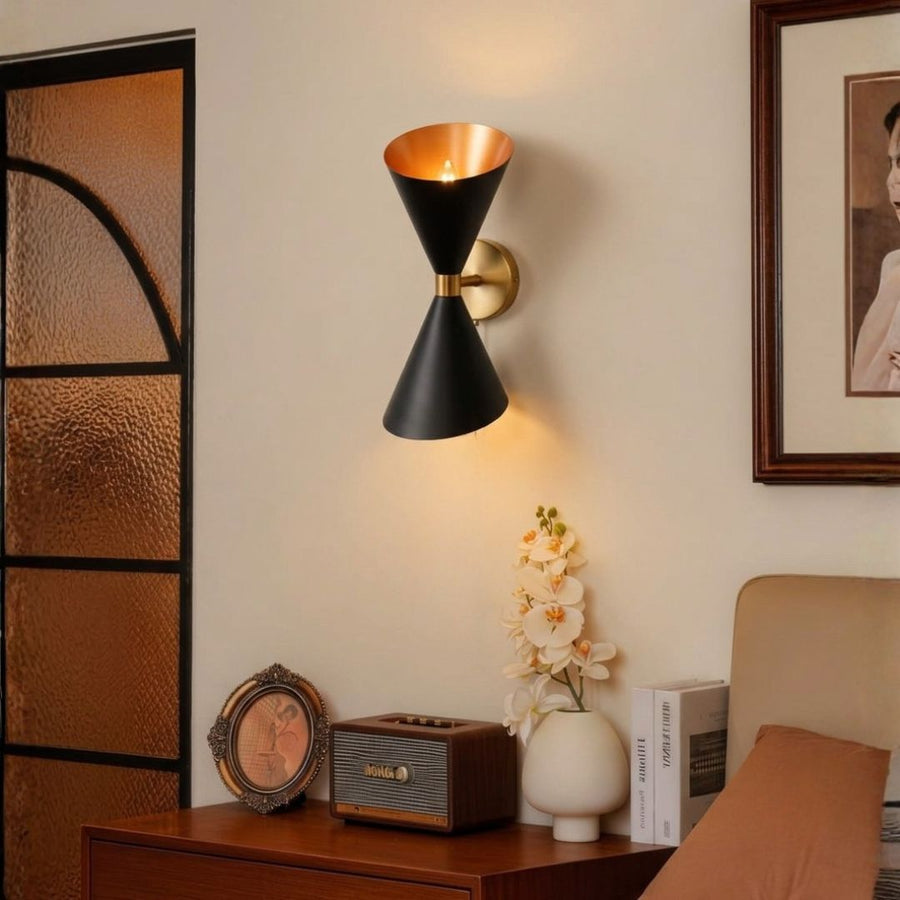 Modern black and gold brass hourglass wall sconce fixture illuminated beautifully in a cozy bedroom setup corner.