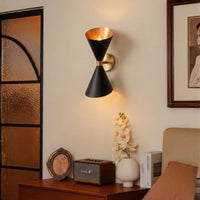Modern black and gold brass hourglass wall sconce fixture illuminated beautifully in a cozy bedroom setup corner.