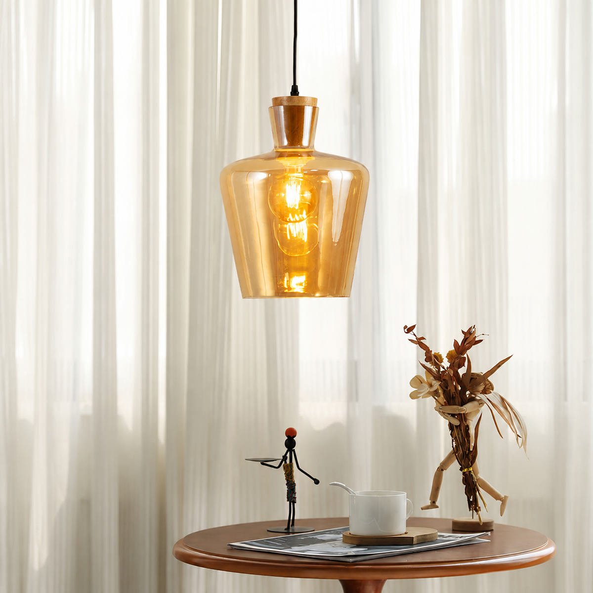 A warm amber glass pendant light hangs over a small round wooden table in a softly lit room with sheer curtains. The glowing bulb is visible inside the translucent amber shade, and decorative objects sit on the table below.