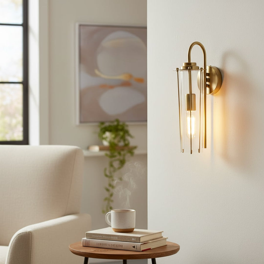 A modern gold wall light with a tall glass shade glowing warmly beside a cozy armchair creating a soft ambient look in a calm living room scene.