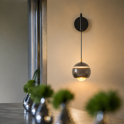 A modern wall-mounted pendant light with a dark round backplate, thin black cord, and a metallic gray glass globe shade glowing warm yellow, shown in a softly lit interior setting.