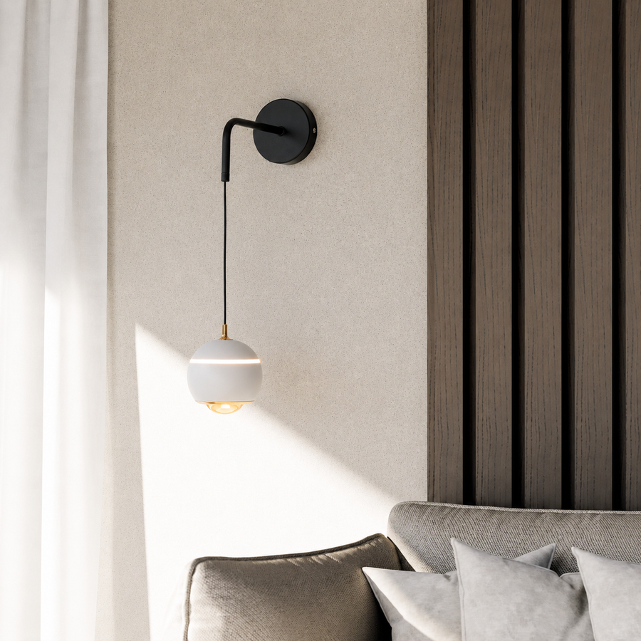 A modern wall-mounted sconce with a black metal arm and a white spherical shade, shown lit in a styled interior next to a sofa and textured wall paneling.