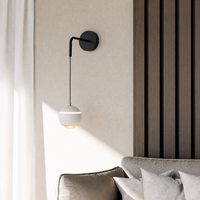 A modern wall-mounted sconce with a black metal arm and a white spherical shade, shown lit in a styled interior next to a sofa and textured wall paneling.