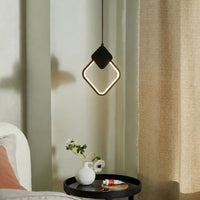 A modern diamond-shaped pendant light with a glowing white LED inner ring and a black outer frame, hanging in a styled bedroom or living space next to a beige curtain and above a small black side table.
