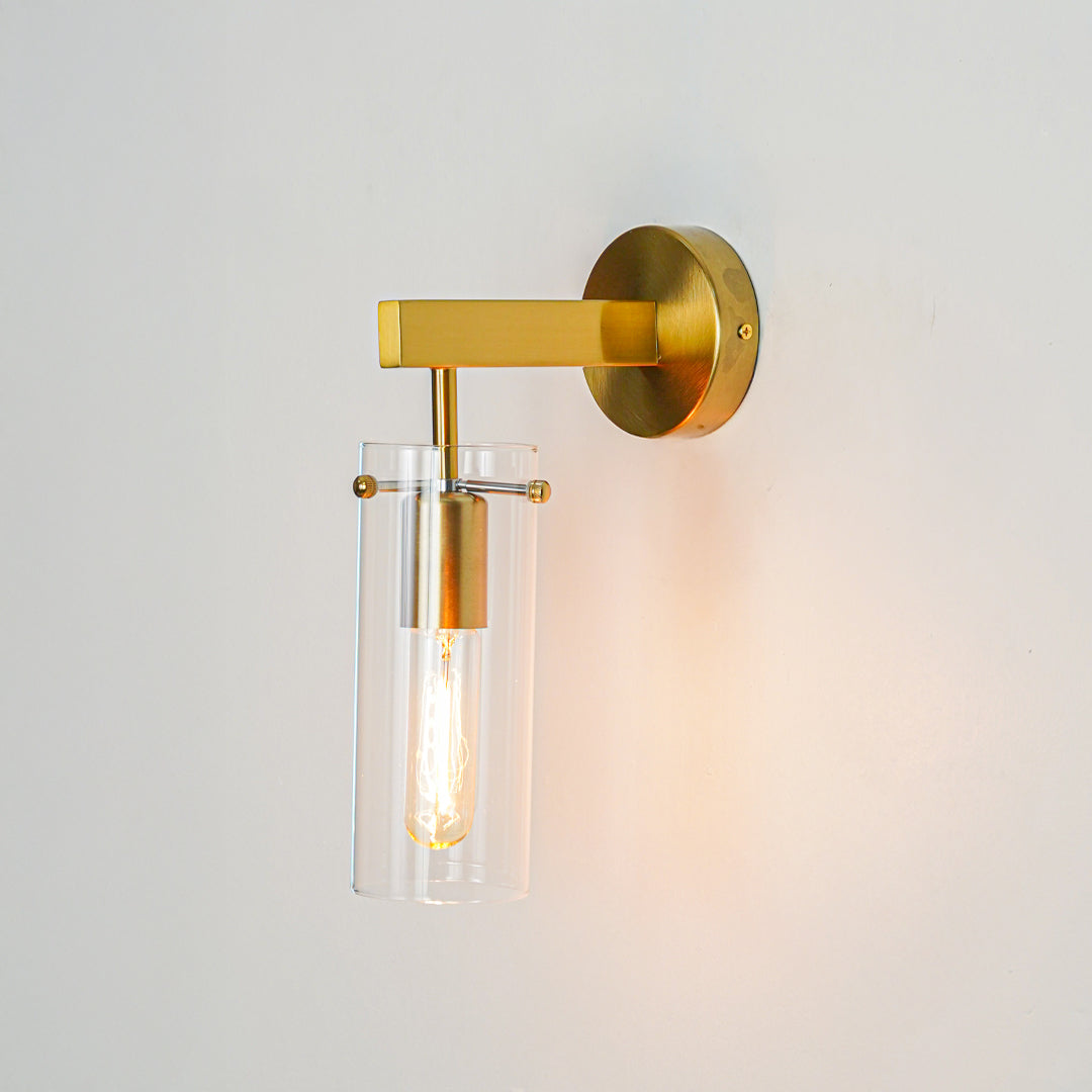 Modern gold metal wall light with cylindrical glass shade and exposed filament bulb mounted on clean white wall background for luxury interior lighting.