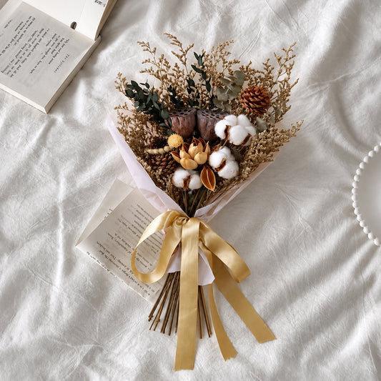 A dried flower bouquet with cotton bolls, pinecones, and a gold ribbon, arranged on white fabric with an open book nearby.