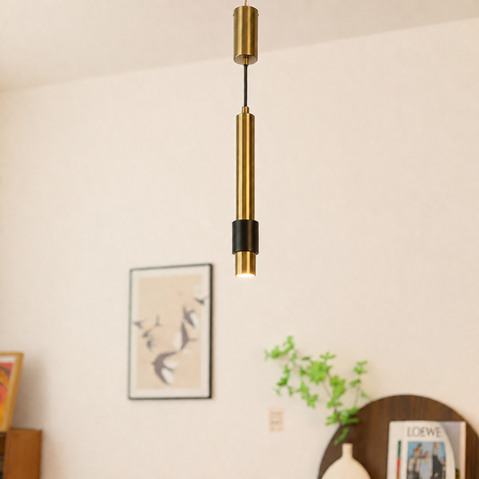 A modern linear pendant light with a brushed gold finish and a black accent band, hanging in a minimalist room with a warm off-white wall and framed art.