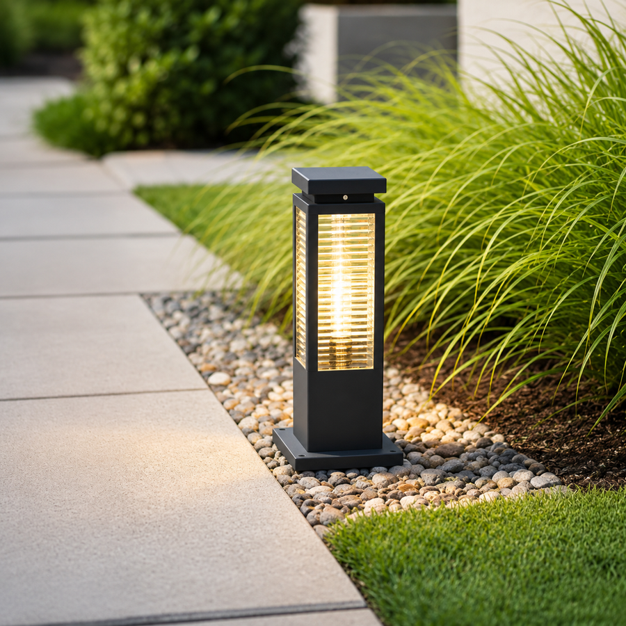 A modern outdoor bollard light with a black metal frame and warm glowing glass, positioned on a bed of small white gravel beside a concrete walkway and green ornamental grass.
