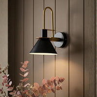 A modern wall sconce with a black conical shade, a gold-toned arched arm, and a round black-and-white wall plate, mounted on a brown wood-paneled wall with warm light shining downward. Pink dried flowers appear in the lower left corner.