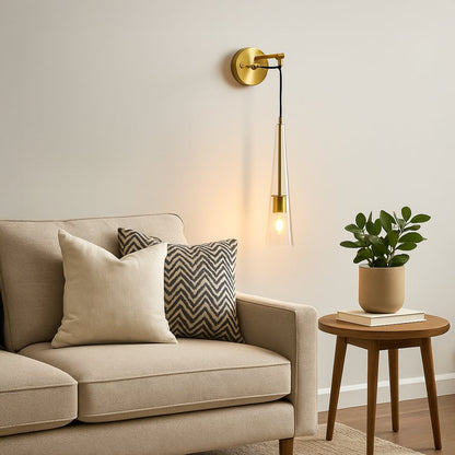 Modern gold wall sconce with hanging tapered glass shade illuminated above beige sofa in living room creating cozy warm reading light.