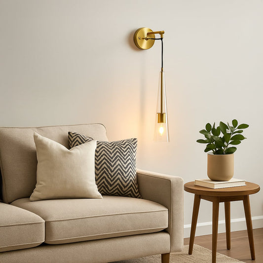 Modern gold wall sconce with hanging tapered glass shade illuminated above beige sofa in living room creating cozy warm reading light.