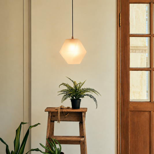A modern hexagonal pendant light with a warm white frosted glass shade hangs above a small wooden stool holding a potted fern in a styled interior setting.