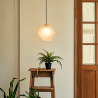 A modern hexagonal pendant light with a warm white frosted glass shade hangs above a small wooden stool holding a potted fern in a styled interior setting.