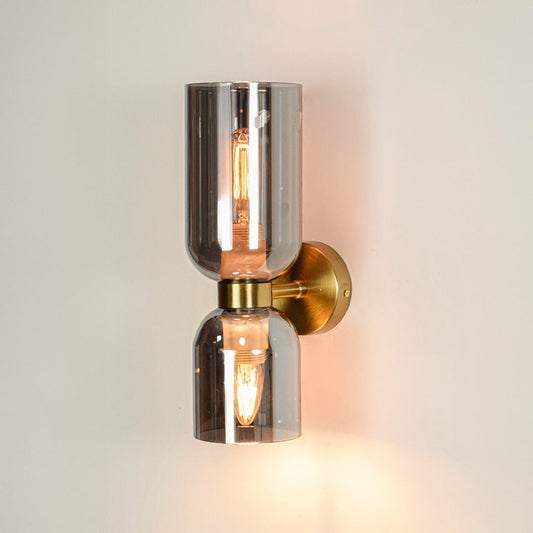 Gold metal wall light illuminated with warm bulb inside smoked glass shades on white background.