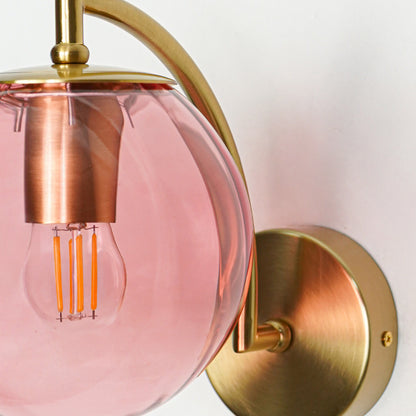 Close up view of blush pink glass wall sconce with exposed filament bulb and gold metal mount against white wall background for modern decor.

