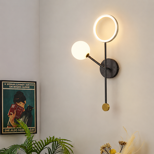 Modern wall sconce with a circular ring light and a spherical globe bulb on a black metal arm, mounted on a light beige wall in a styled interior setting.