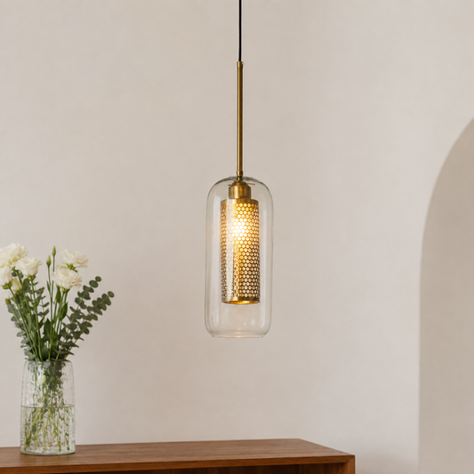 A modern pendant light with a clear glass cylindrical shade and a brass inner mesh diffuser, hanging above a wooden console table with a vase of white flowers against a warm off-white wall.