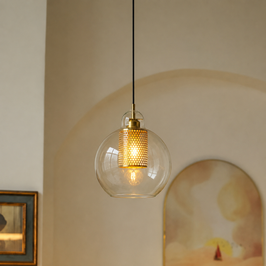 A modern pendant light with a clear glass globe shade, a gold-toned perforated cylindrical inner shade, and a black hanging cord, shown lit in a softly styled interior setting.