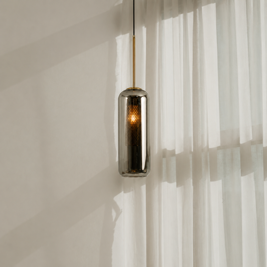 A modern cylindrical pendant light with a brushed metallic finish and a warm glowing LED inside, hanging in front of sheer white curtains.