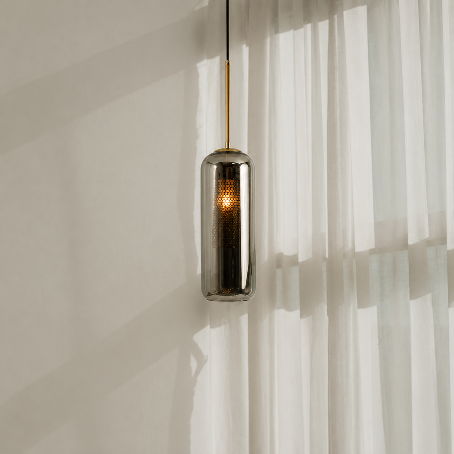 A modern cylindrical pendant light with a brushed metallic finish and a warm glowing LED inside, hanging in front of sheer white curtains.