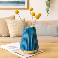 Contemporary blue vase with a pleated design and metallic gold base arranged on a coffee table with bright yellow blooms complementing cozy beige cushions in a minimal living space.