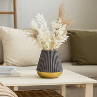 Grey ribbed ceramic vase with a golden base filled with dried pampas grass and white foliage placed on a wooden coffee table in a cozy living room with neutral-toned cushions and soft textures.