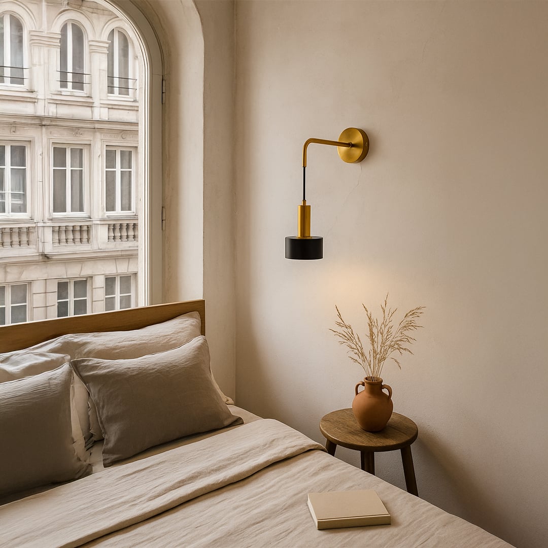 Modern gold wall sconce with hanging black cylinder shade illuminated next to bed in cream bedroom illustrating cozy reading light.