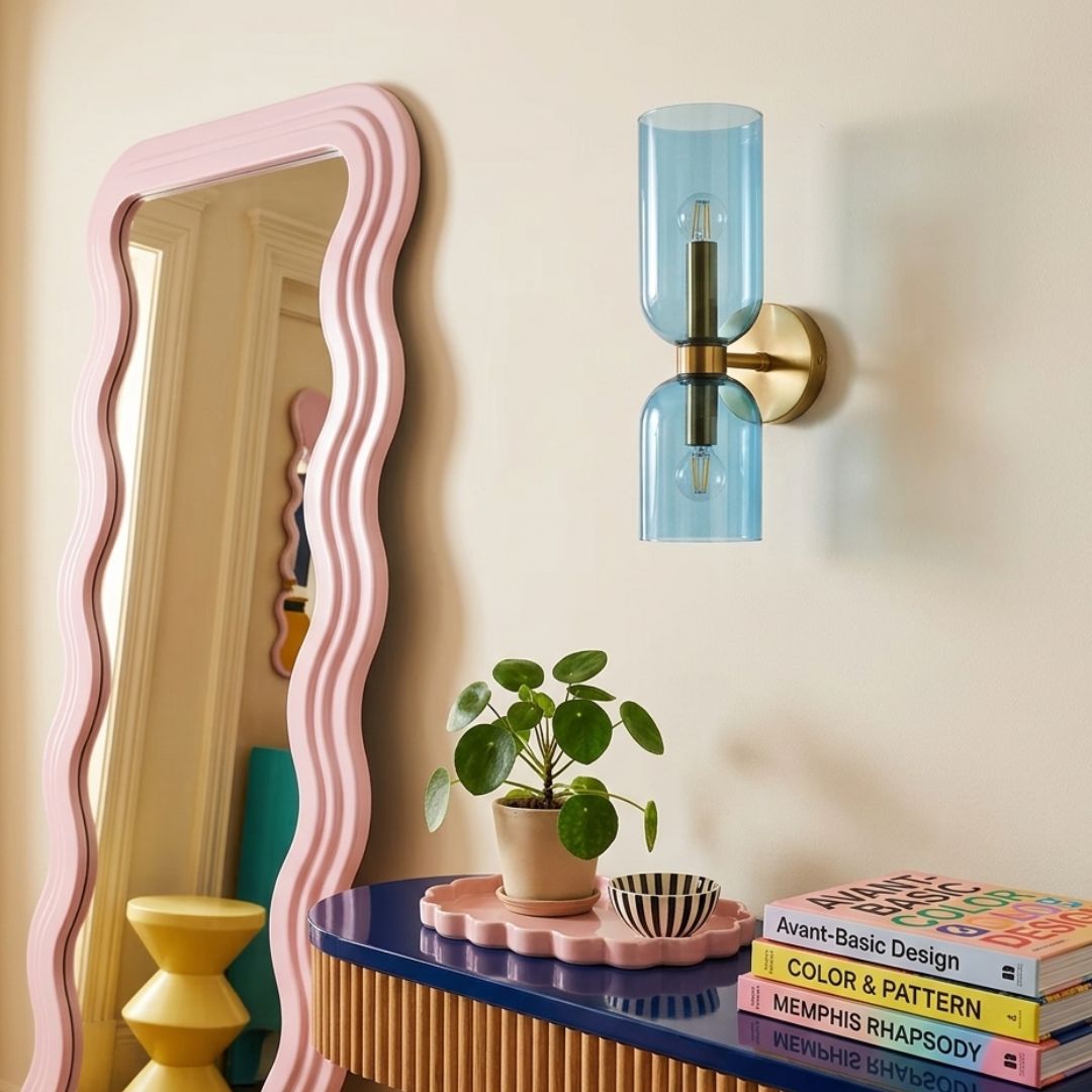 A modern wall-mounted light fixture with a translucent light blue glass cylinder and a brass-toned metal mount, shown on a beige wall above a styled console table with a plant and books.