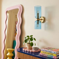 A modern wall-mounted light fixture with a translucent light blue glass cylinder and a brass-toned metal mount, shown on a beige wall above a styled console table with a plant and books.
