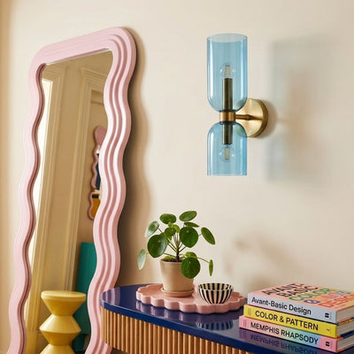 A modern wall-mounted light fixture with a translucent light blue glass cylinder and a brass-toned metal mount, shown on a beige wall above a styled console table with a plant and books.