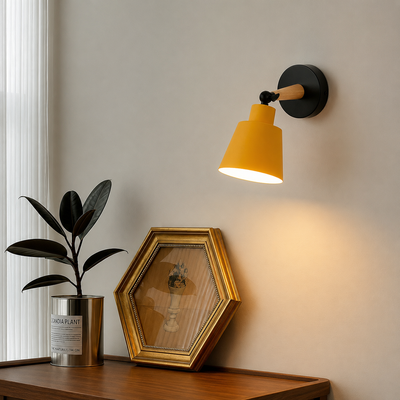A modern wall-mounted sconce lamp with a bright yellow cone shade, black wall and joint hardware, and a light wood accent, shown lit on a beige wall above a wooden console with a framed picture and potted plant.