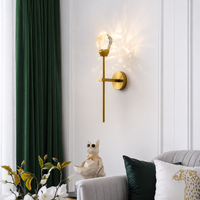 A modern brass wall sconce with a faceted crystal-like globe bulb, mounted on a white paneled wall next to a dark green curtain, above a light gray sofa with decorative pillows.