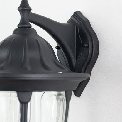 Closeup of black downward wall lantern showing curved glass panels and decorative finial with traditional coach light design.