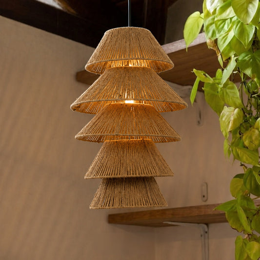 A tiered pendant light made from natural woven raffia or jute, hanging in a warm indoor setting with a beige wall, wooden shelf, and green trailing plant visible on the right.