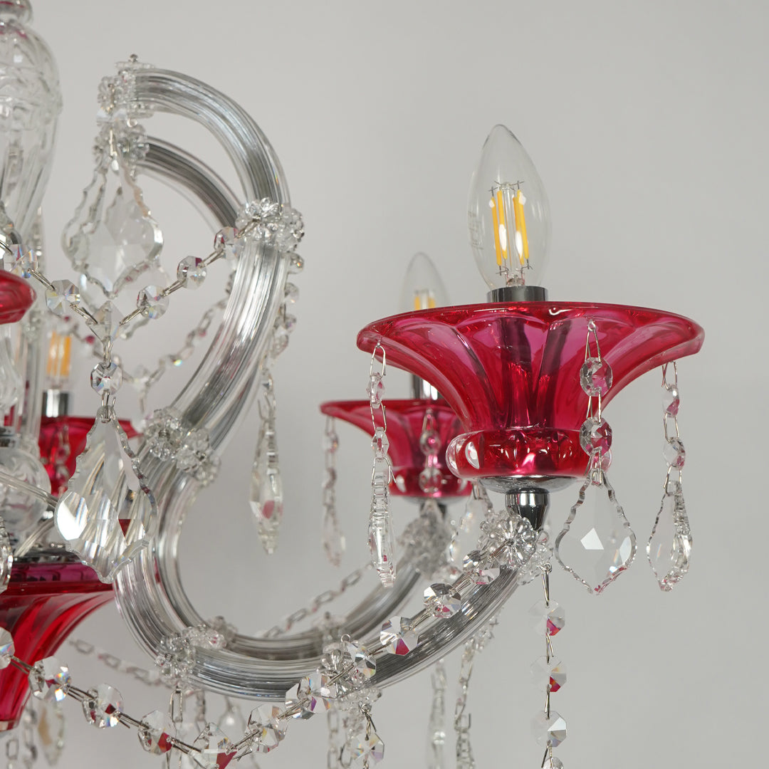 A close-up of a crystal chandelier featuring red glass candle-style shades, clear glass arms, and hanging teardrop crystal pendants against a light gray background.