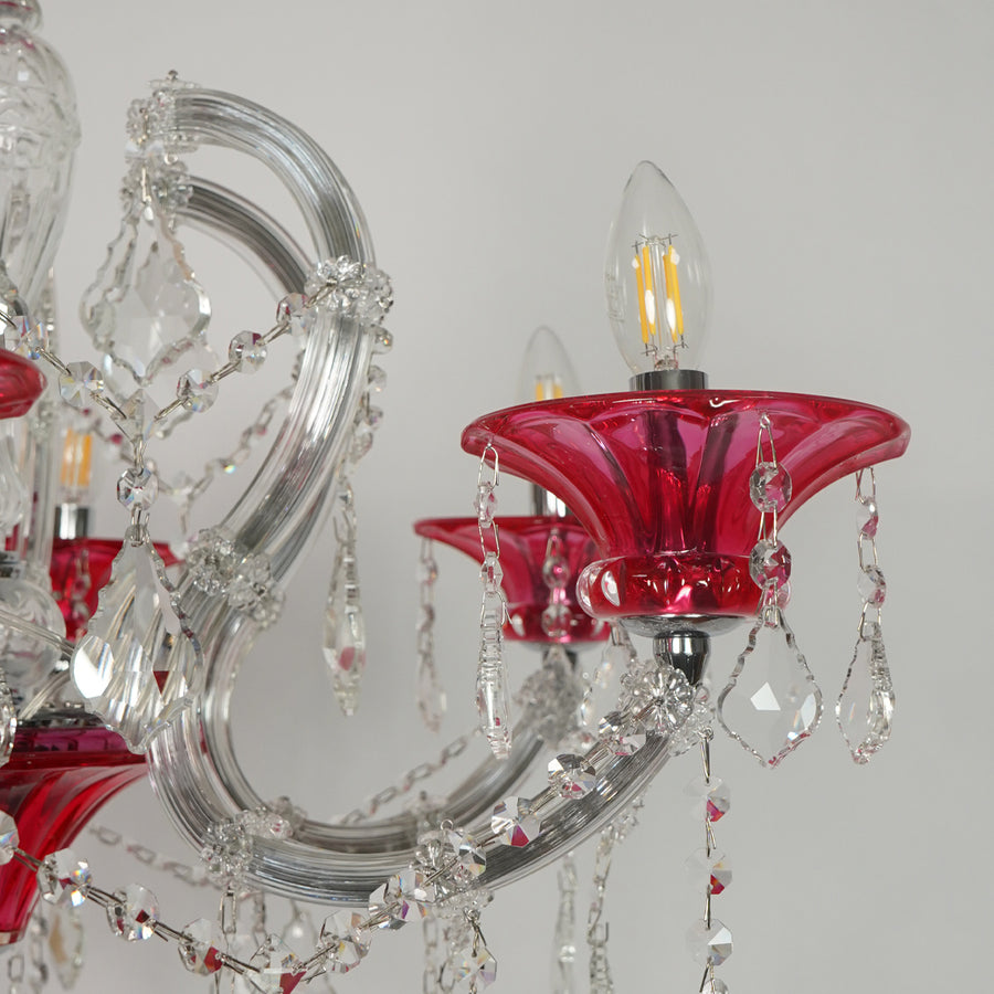 A close-up of a crystal chandelier featuring red glass candle-style shades, clear glass arms, and hanging teardrop crystal pendants against a light gray background.