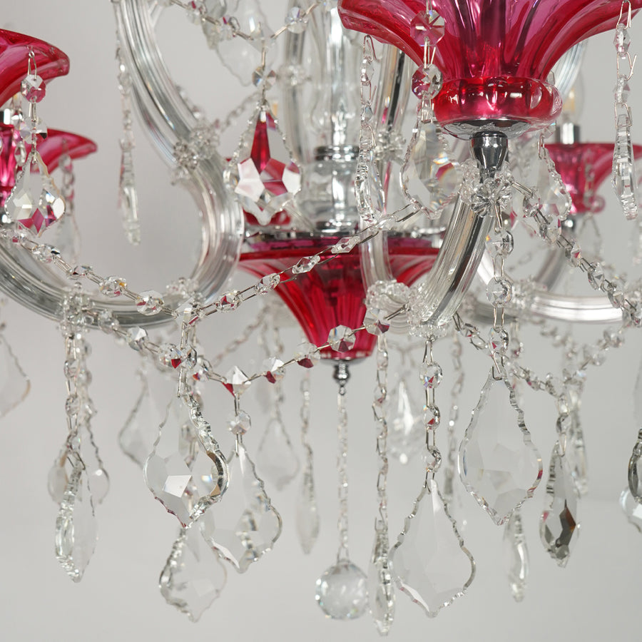 A close-up of a crystal chandelier with vibrant pink glass shades, clear faceted crystal teardrop pendants, and a silver-toned metal frame.