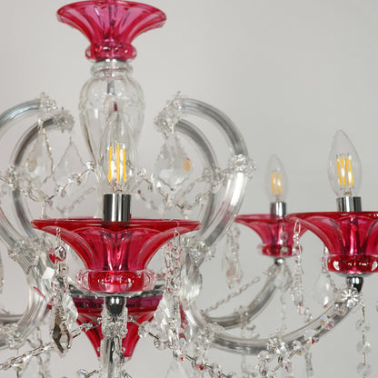 A close-up of a crystal chandelier featuring vibrant red glass cup holders and clear glass arms with hanging crystal prisms, shown against a neutral light gray background.