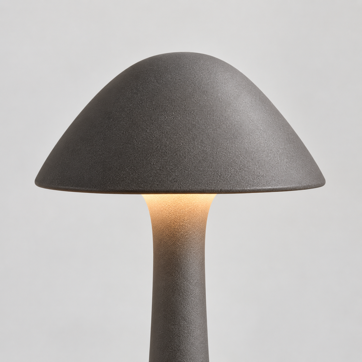 A mushroom-shaped table lamp with a textured dark gray ceramic shade and stem, glowing with a warm light from the center, set against a neutral light gray background.