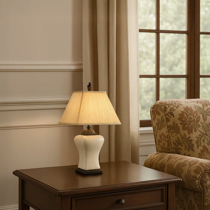 Cream shade beige ceramic table lamp on dark wooden side table with light on beside upholstered chair for warm traditional décor.