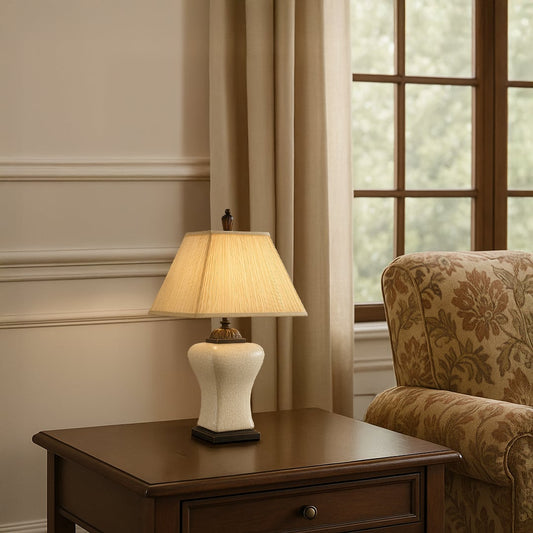 Cream shade beige ceramic table lamp on dark wooden side table with light on beside upholstered chair for warm traditional décor.